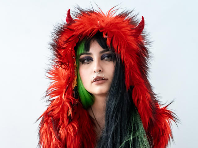 AngelBlakee with bright red, fluffy hood and black-green hair, nose ring, and green ear, wearing makeup, looking serious, upper body shot, white background.