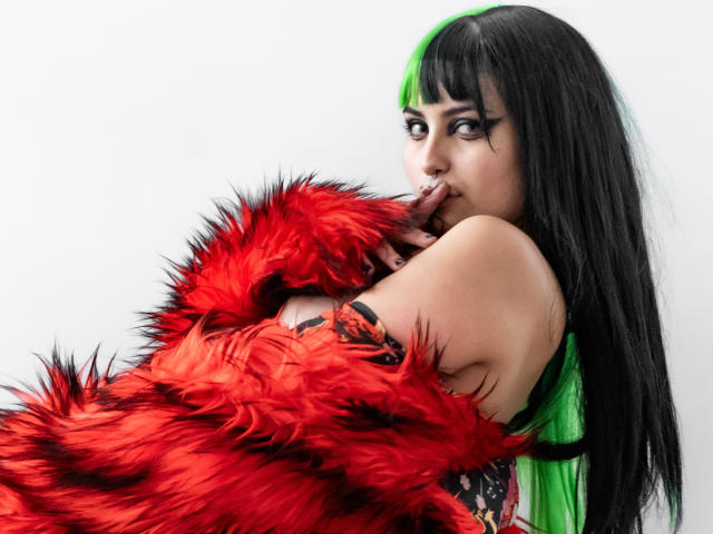 AngelBlakee has black, straight hair with a bright green streak. She wears a red, fluffy fur coat with black stripes, partially covering her shoulders. She looks over her shoulder, biting her finger with a sultry expression. Her makeup is bold, with thick black eyeliner and dark eyeshadow. She has a fair skin tone and a tattoo on her right arm. The image is sharp, with a clean white background. The contrast between the red fur and her green hair is striking.