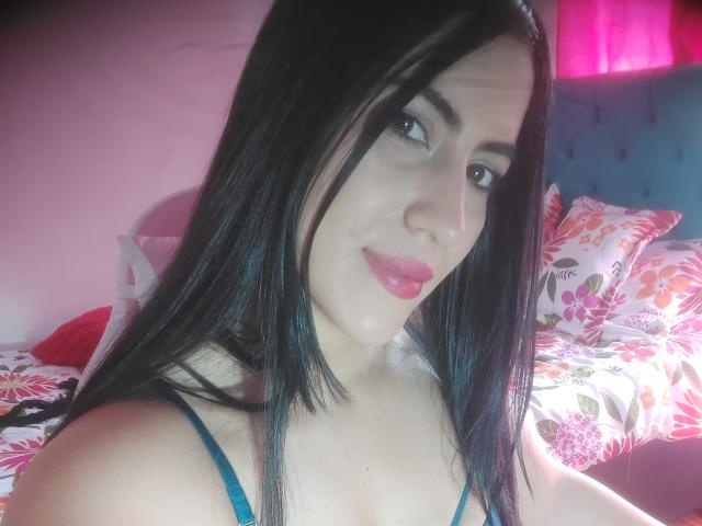 SofiaFountaine with black hair, pink lipstick, blue bra, and light skin, smiling at the camera, showing her tits in a bright, colorful bedroom.