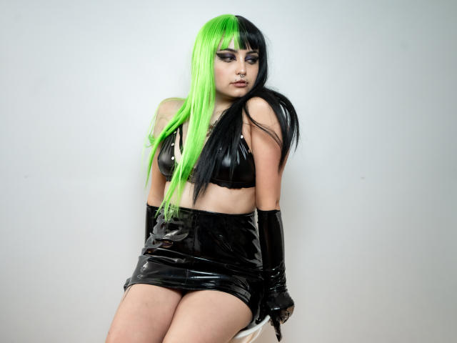 AngelBlakee with green and black half-up ponytail, black latex crop top and skirt, black gloves, sitting, showing her ass, looking down with a serious expression.