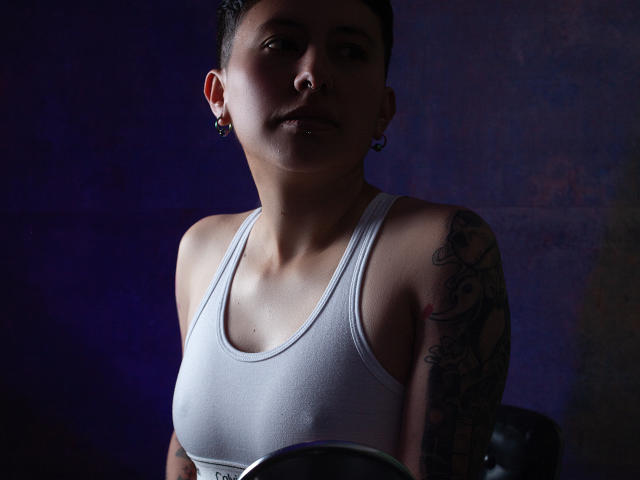 GabiAndMei with short dark hair, white tank top, tattoo on right arm, earrings, nose ring, holding a small round mirror, dim blue-purple light.
