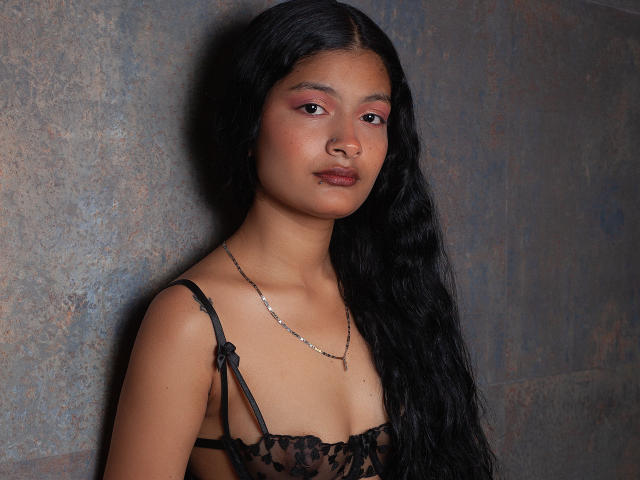 GabiAndMei with long black hair, brown skin, pink eyeshadow, black lace lingerie, small tits, and a silver necklace, standing against a textured wall.