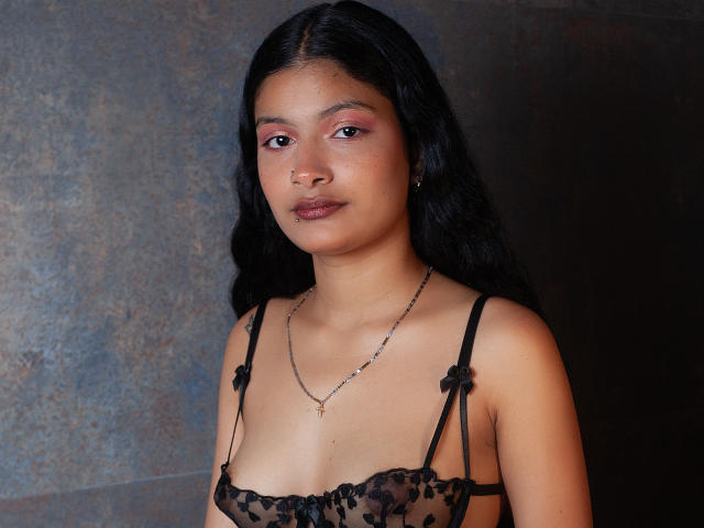 GabiAndMei with brown skin, black hair, and pink eye makeup, wearing a black lace bra, necklace, and earrings, looking slightly to the right. Boobs are visible.