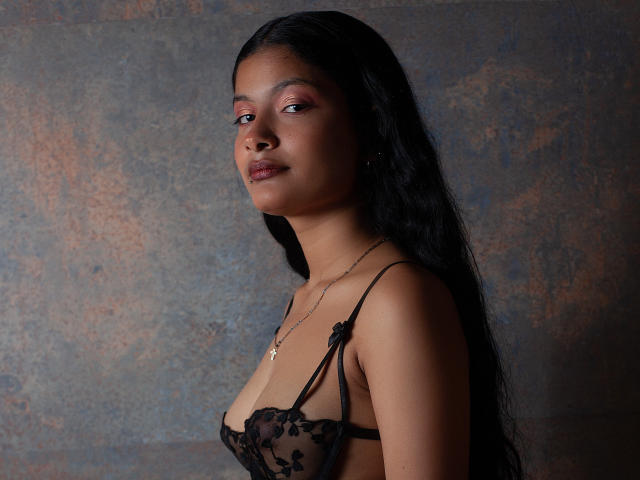 GabiAndMei with long black hair, dark skin, and pink eyeshadow, wearing a black lace bra, showing her small tits and a cross necklace, looking slightly to the side.
