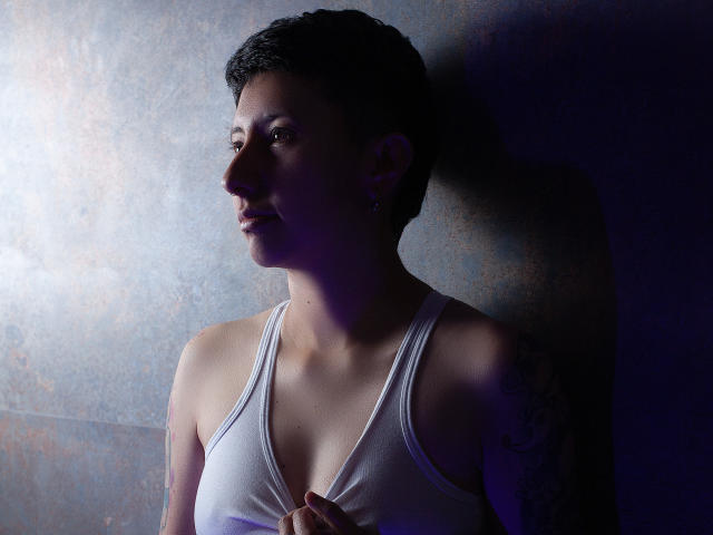GabiAndMei with short black hair, white tank top, small breasts, tattoo on right arm, holding shirt, looking to the side, shadowed, purple light, textured wall, slight smile.