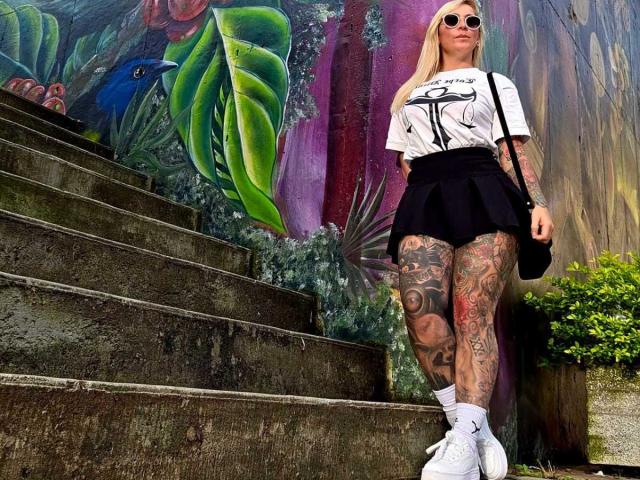 SaraValkyria stands on concrete stairs, wearing a white tee, black skirt, white socks, and white sneakers, showcasing tattooed thighs and wearing round sunglasses.