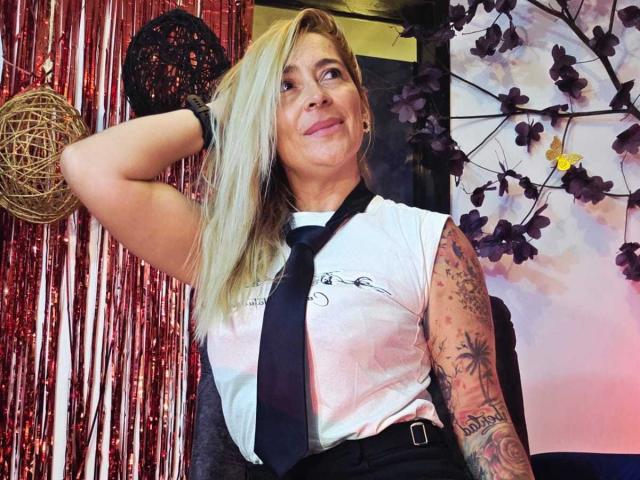 SaraValkyria with blonde hair, black tie, white sleeveless shirt, tattooed arm, smiling, hand in hair, red and silver background, purple flowers. Boobs are not visible.