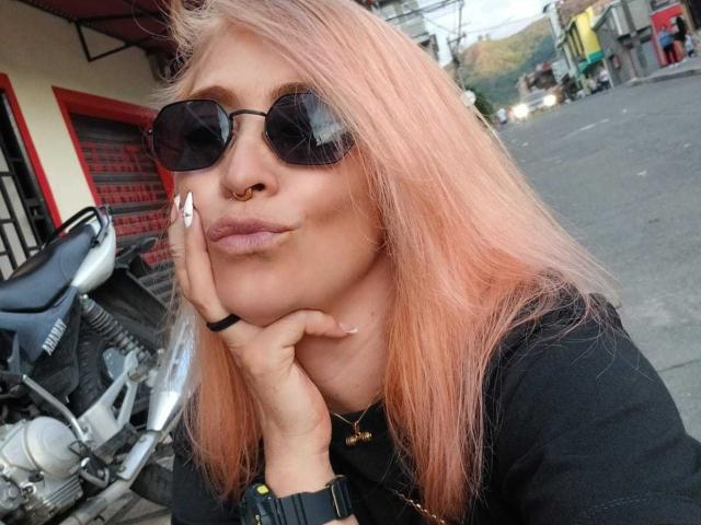 SaraValkyria with pink hair, black sunglasses, nose ring, and pouty lips, resting her cheek on her hand, wearing a black shirt and black watch, next to a motorcycle.