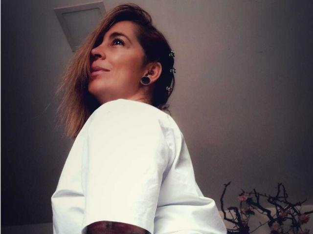 SaraValkyria with blonde hair, white shirt, black ear gauges, looking up, smiling, visible upper back, small tattoo on right arm, medium-sized breasts.