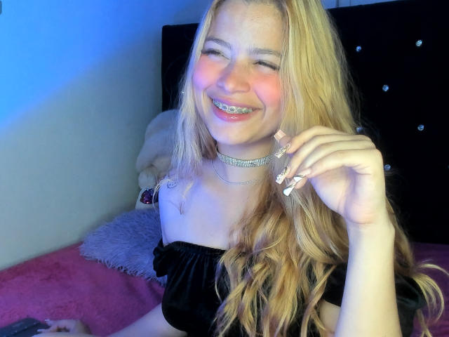ShantalSummer with long blonde hair, smiling with teeth showing, wearing a black off-shoulder top and sparkly choker, touching her cheek. Her ass is partially visible.