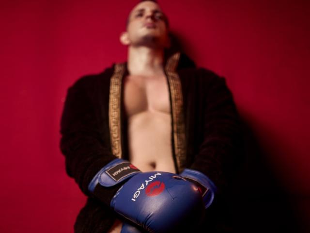 AlessandroVieri stands with a blurry red background, wearing a dark robe open to show a bare chest, and blue boxing gloves, looking up with a slight smile.