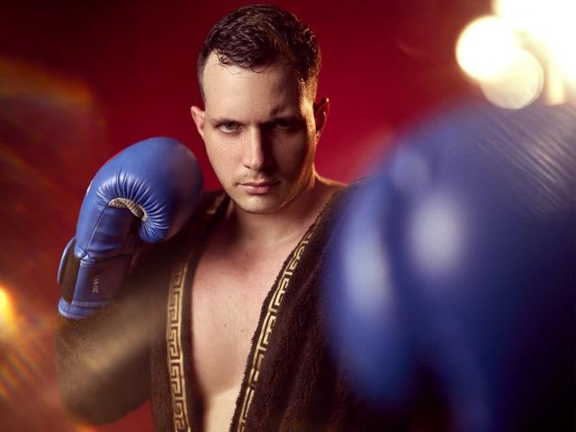 AlessandroVieri, shirtless, wearing blue boxing gloves, brown robe with gold trim, intense look, right fist raised, blurry blue fists in foreground, short dark hair, red background.