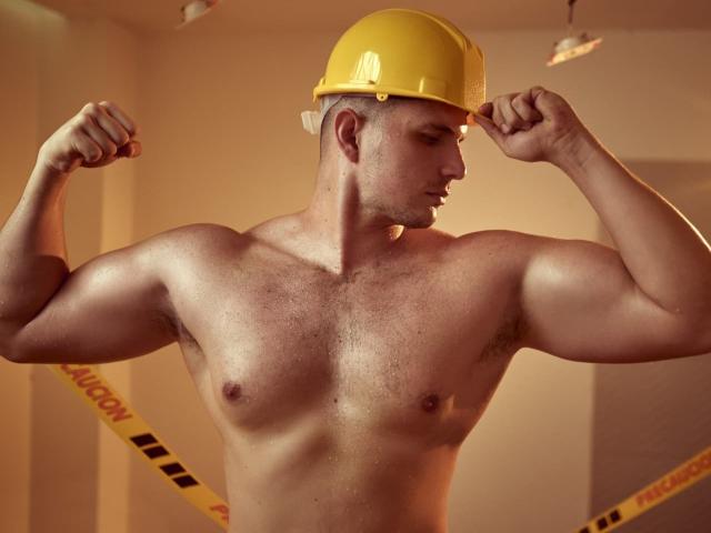 AlessandroVieri, shirtless with a yellow hard hat, flexes muscular arms, showing defined chest and abs, with a slight tan, in a room with caution tape.