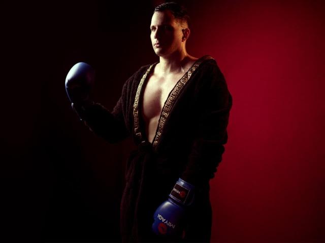 AlessandroVieri, a muscular guy with a deep tan, stands in a dark brown robe, open to show his chest, wearing blue boxing gloves, holding a blue boxing helmet.