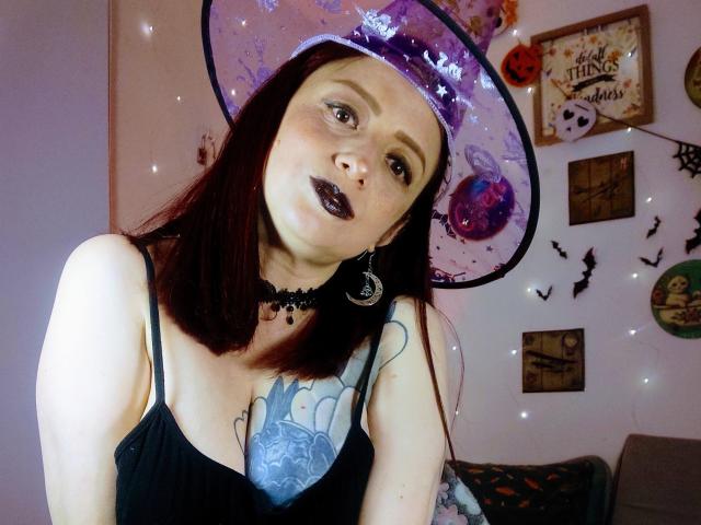 CatalinaMonyque with pale skin, dark lipstick, and red hair, wearing a purple witch hat, black choker, and black dress, showing her tits and tattoo, in a Halloween-themed setting.