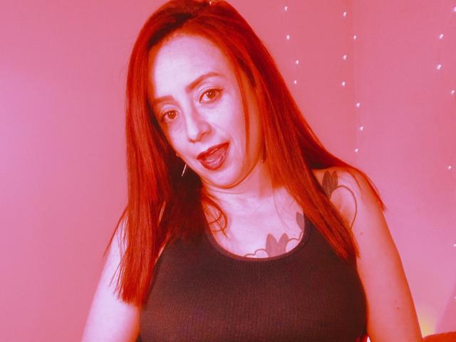 CatalinaMonyque has red hair, a black tank top, and a heart tattoo on her chest. Her tits are medium-sized and she's sticking out her tongue. Red lighting.