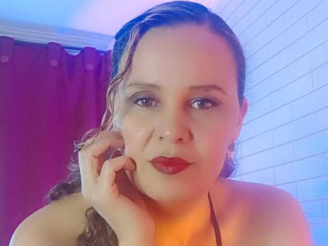 VittoriaI has wet brown hair, red lipstick, and a serious expression, touching her cheek with a hand, wearing a black halter top, in a brightly lit room.