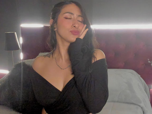 ZaraBeicker with her eyes closed, black off-shoulder top, and hand near her face, showing her tits and a subtle smile, in a dimly lit room.