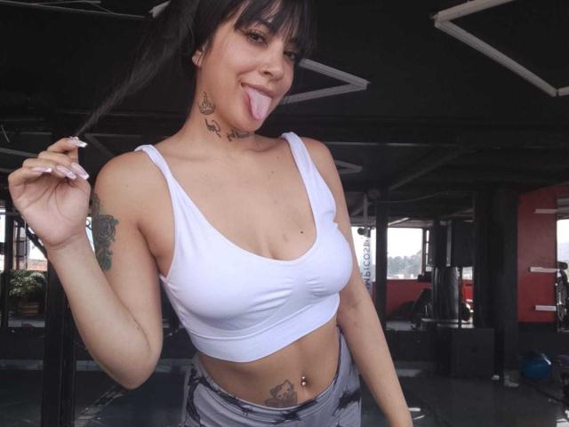 AnalisseJade con le trecce, che fa penzoloni con la lingua, indossa un reggiseno sportivo bianco che mette in mostra il seno e un piercing all'ombelico, con tatuaggi sul collo e sul petto..