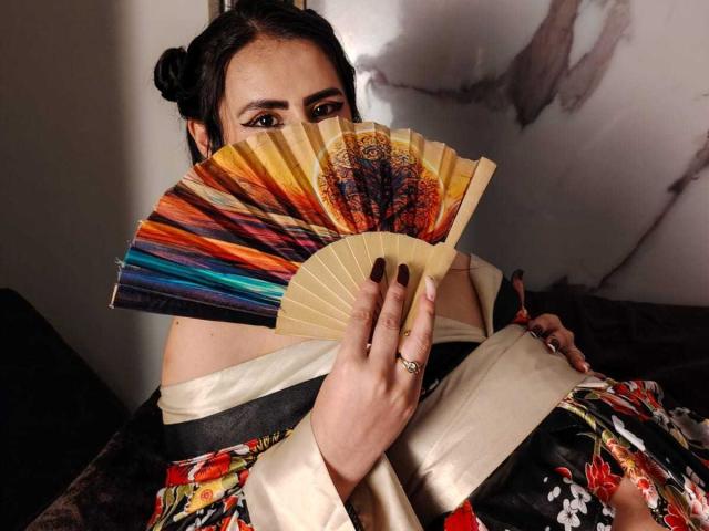 DemiMistral holding a colorful fan with her fingers over her tits, wearing a floral kimono, dark nails, and a ring, with her eyes peeking through.