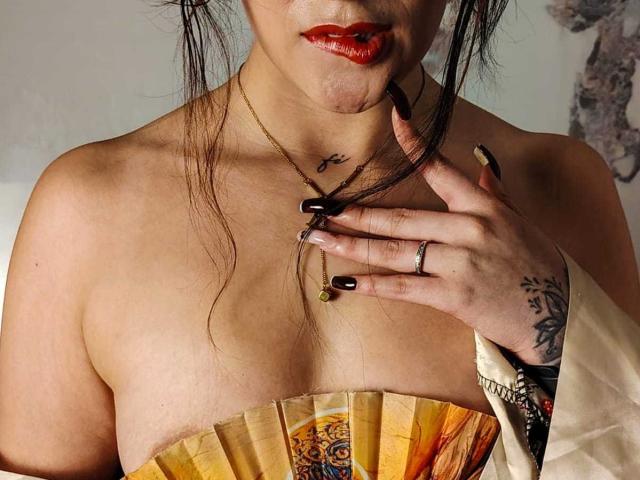 DemiMistral with red lipstick, black nails, gold necklace, ring, and hand on chest, holding fan over her tits, tattoo on neck, tattoo on wrist, dark hair, tan skin.
