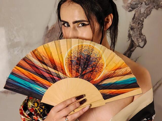 DemiMistral holding a colorful fan with her hand over her tits, dark hair in a ponytail, black eyeliner, beige off-shoulder top, and dark nails.