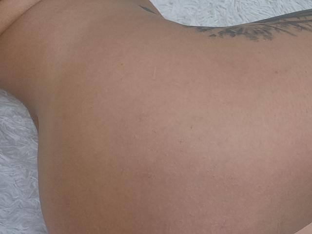 Close-up of TheSunshine's lower back and buttocks. TheSunshine has light brown skin with a smooth texture and subtle stretch marks. A small, dark tattoo of a leafy design is on the right side of TheSunshine's lower back. The background is a white, textured surface. The image focuses on the natural curvature and skin details. The tattoo is slightly angled, adding a subtle dynamic to the composition. The image is sharp with a clear focus on the skin and tattoo.