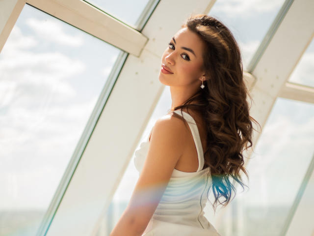 EricaSiin stands in a sunlit room with a glass ceiling, looking over her shoulder. She has long, wavy brown hair with blue tips, and is wearing a white sleeveless dress. Her brown eyes and light pink lips are prominent. Soft light and lens flares highlight her. The image uses natural light, emphasizing her fair skin and subtle makeup. EricaSiin's slightly turned head and relaxed pose create a casual, yet confident vibe.