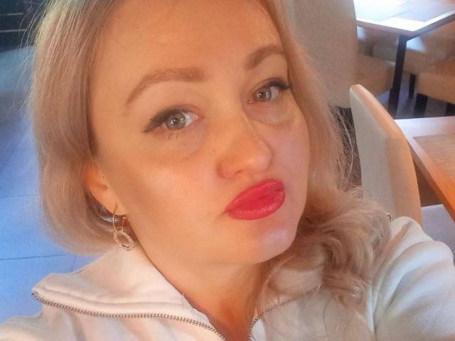 CurvyMuses with blonde hair, pink lipstick, and hoop earrings, wearing a white hoodie, has a pouty expression, blue eyes, and is taking a selfie.