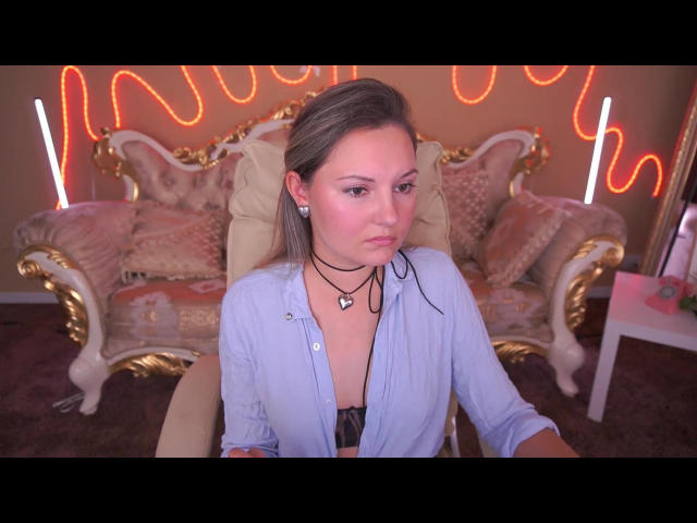 DorisBliss with pouty face, wearing unbuttoned blue shirt, black choker, showing small tits, sitting in ornate chair, neon orange lights in background, white table to right.