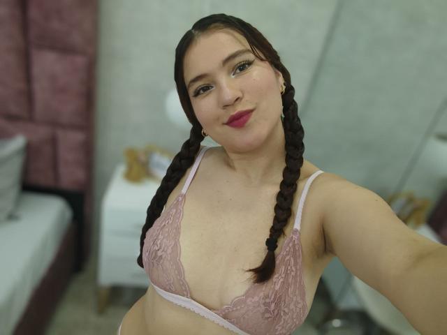 CelesteDash with braided hair, pink lace bra, and red lipstick, showing her tits in a selfie. She's smiling and has light skin and dark eyes.