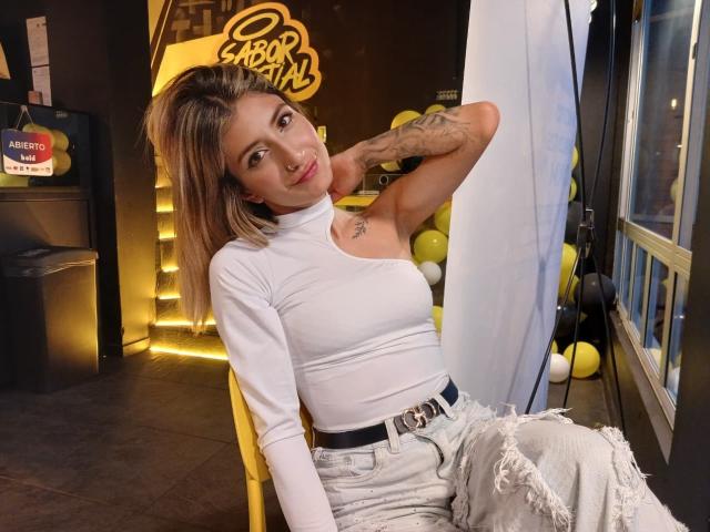 Kaiix with blonde hair, white turtleneck, and ripped jeans, sitting on a yellow chair, arm raised, showing tattoos, and a black belt with a gold buckle.