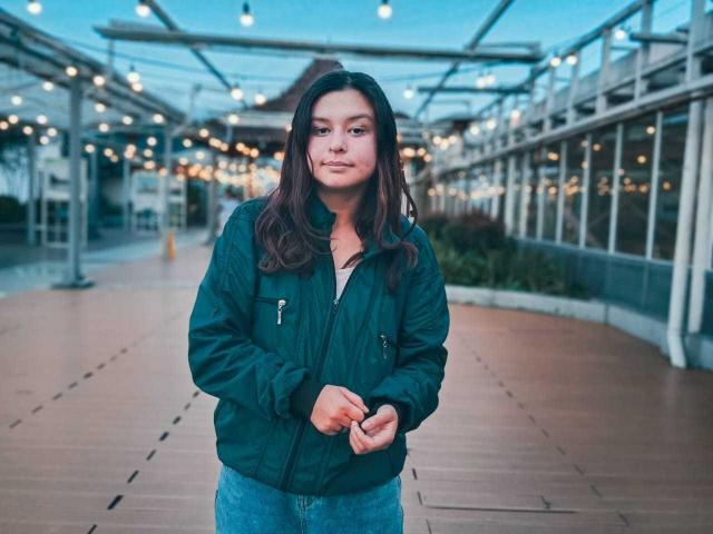 DyanaMoon stands in a blue-green jacket with zippers, hands clasped, looking forward. She's got brown hair and a subtle smile. She's wearing blue jeans, in a twilight setting.