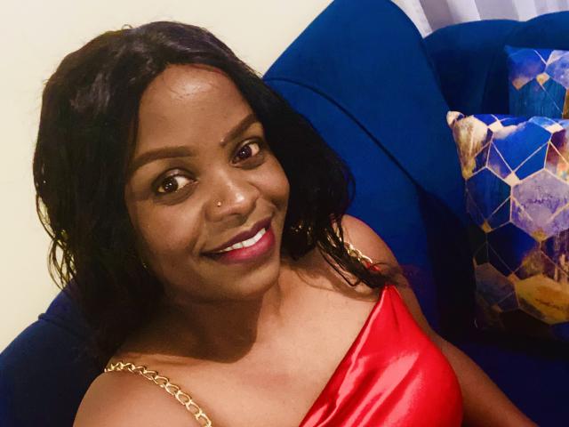 Choccoo has dark skin, black wavy hair, brown eyes, and a nose ring. She's wearing a red satin top with gold straps, smiling, and sitting on a blue couch.
