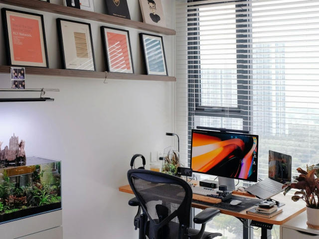 Modern office setup with a desk in the foreground. A black mesh office chair faces a flat-screen monitor with a colorful abstract wallpaper. Shelves above hold framed art and small photos. To the left, a lit aquarium with green plants and fish. Right window with white blinds. Desk has a black lamp, a potted plant, and a laptop. rs0307Test1234-hot is not present. Clean, bright, and organized.