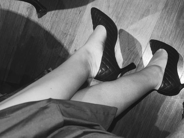 Black-and-white photo of AmelineRousseau's legs in dark, textured high heels on a wooden floor. The heels have a sleek, glossy finish and sharp, pointed toes. AmelineRousseau's legs are crossed at the ankle, with her left leg slightly in front of the right. The wooden floor has a subtle grain pattern, and a shadow is cast from the heels. The image emphasizes the contrast between the dark heels and the lighter skin.