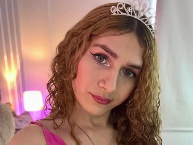 AndyDupont with curly brown hair, wearing a silver tiara, pink eyeliner, and pink lipstick, in a pink top, showing a cute face and a small nose ring.