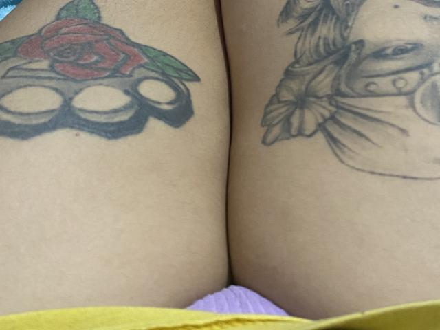 GipsyZ's thighs, showing a red rose tattoo with green leaves on the left and a black-and-grey floral tattoo on the right, with yellow underwear at the bottom.