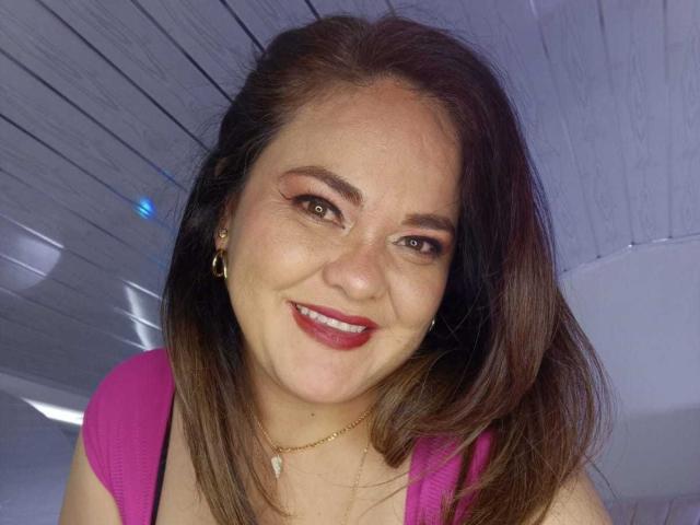 NatashaBlade has brown hair, red lipstick, and is smiling with pink clothes, showing her tits, and wearing a gold necklace in a selfie with white wooden ceiling.