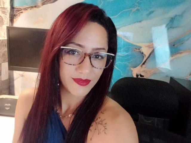 SamanthaCorine with red and black hair, wearing glasses, red lipstick, and a blue halter top, showing a tattoo on her right shoulder, sitting in a black chair.