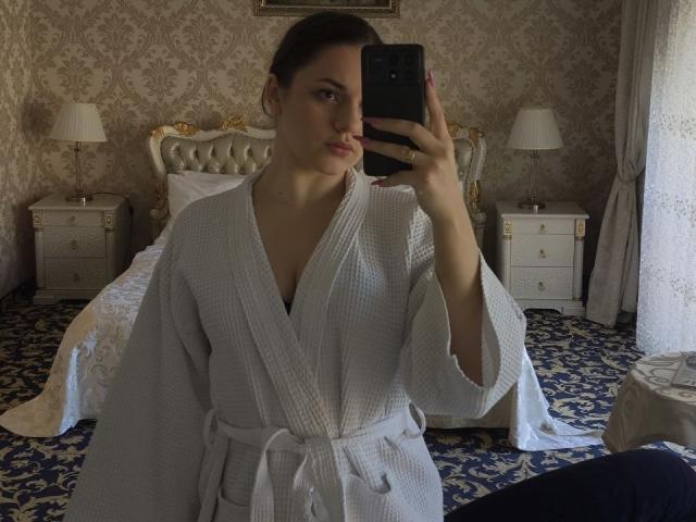 Dreia, com seios pequenos, vestindo um roupão de algodão branco, tira uma selfie no espelho em um quarto luxuoso, segurando um smartphone preto, com cabelo castanho escuro preso em um coque..
