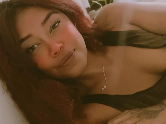 DalilaHeinz, avec ses cheveux roux bouclés, sa peau brune et son maquillage discret, allongée, portant une robe noire à bretelles, un petit collier en argent, les lèvres légèrement entrouvertes et un sourire doux et séduisant..