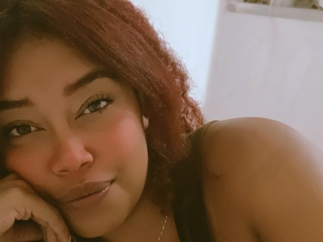 Dalila Heinz, con cabello rojo rizado, ojos verdes y una ligera sonrisa, apoyando su barbilla en la mano, mostrando su hombro descubierto y un pequeño tatuaje. Luz suave en su rostro..