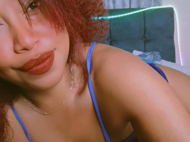 DalilaHeinz with curly red hair, wearing a blue bra, has full red lips and a cute smile. Her brown skin is smooth, showing her small, perky tits.