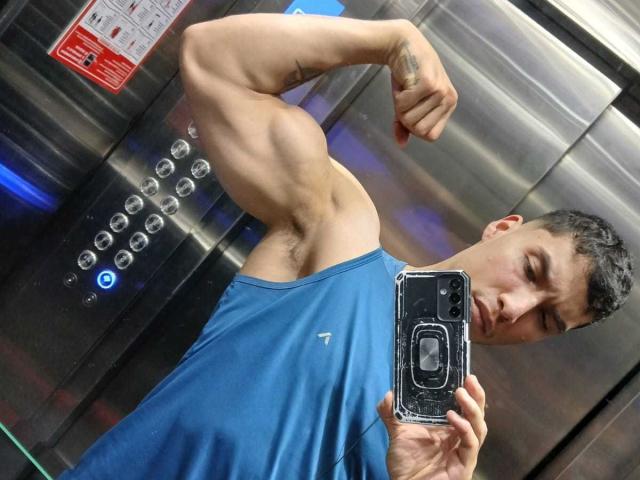 JuanJsmith flexing his muscular arm in a blue tank top, showing a hairy armpit, taking a mirror selfie with a phone in a metallic elevator.