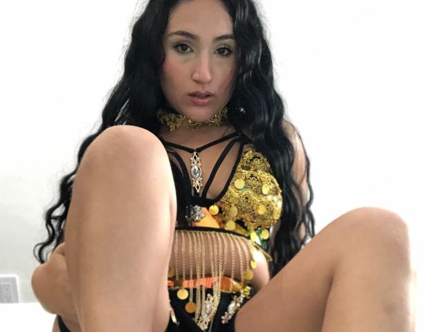 HiilaryDolce with long black hair, wearing a gold and black sequined top, sitting with legs spread, showing her pussy, and a gold choker, looking directly at the camera.