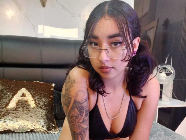AryVixen with dark hair, glasses, and a black bikini top, showing her tattooed arm and cleavage, sitting on a bed with a gold sequin pillow.