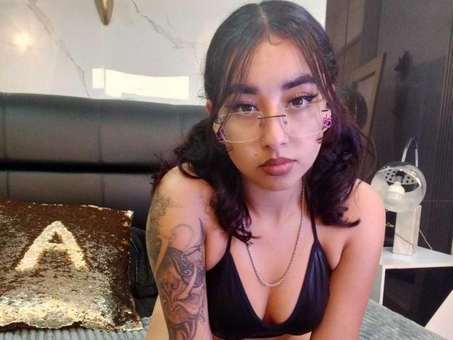 AryVixen with glasses, black hair, and a tattooed arm, wearing a black bikini top, looking at the camera with pouty lips, sitting on a bed with a gold pillow.