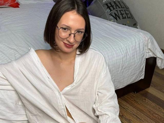 Alliana, con gli occhiali, capelli castani corti, sorridente, indossa una camicia bianca, ampia e con bottoni, seduta sul pavimento di legno di fronte a un letto con lenzuola bianche a righe..