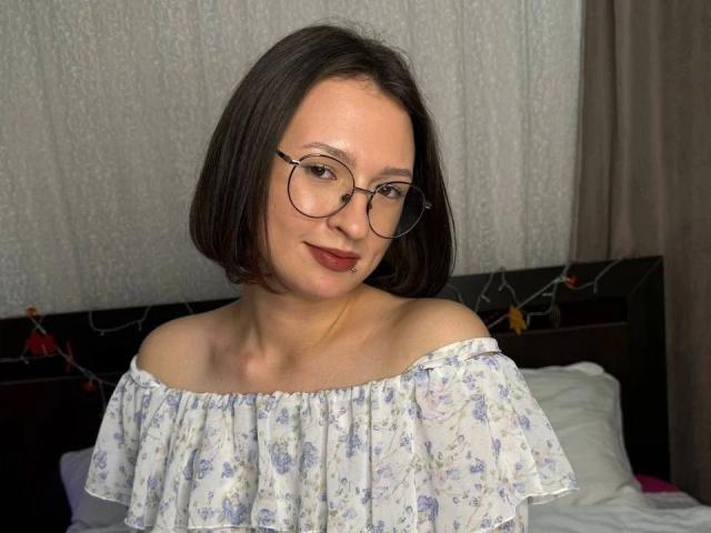 Alliana-hot with short brown hair, glasses, and red lipstick, wearing an off-shoulder floral dress, smiling softly. Boobs partially visible, white fabric background, dark headboard.